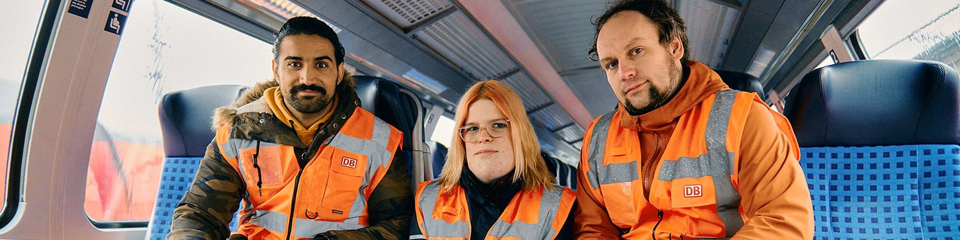 Zwei Männer und in der Mitte eine Frau sitzen in einem Regionalzug, sie tragen Warnwesten.