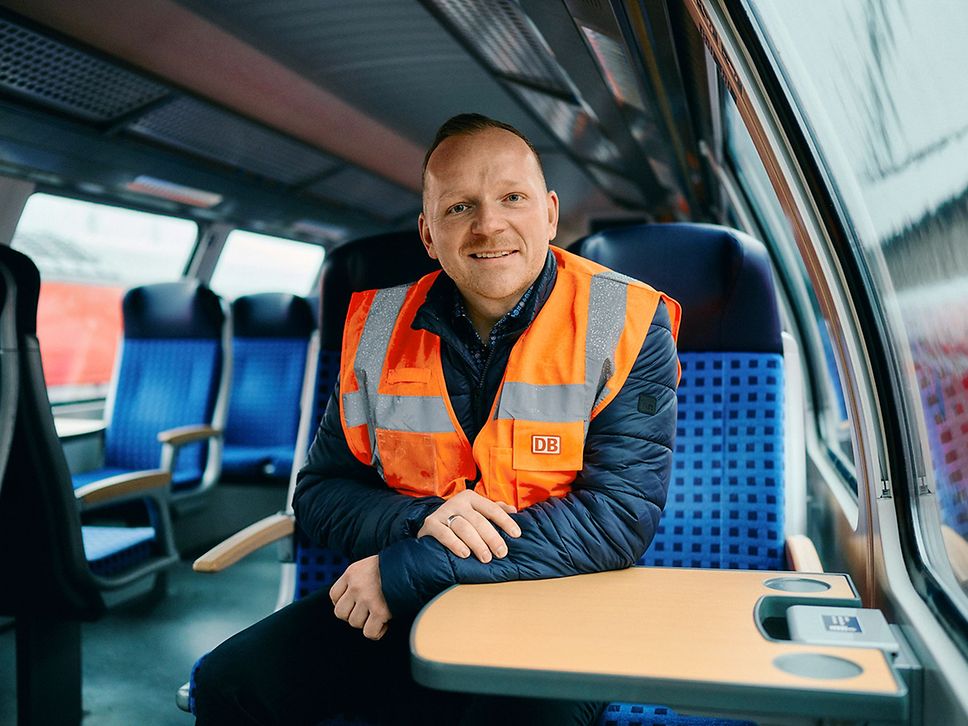 Ein Mann sitzt am Tisch im Regionalzug und lächelt in die Kamera, er trägt eine Warnweste.