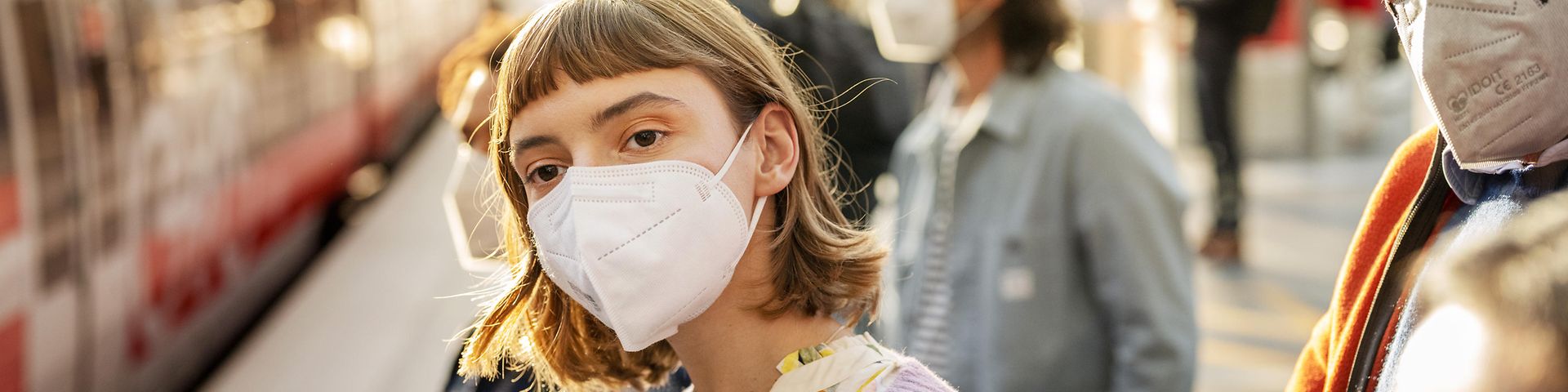 Eine junge Frau mit Maske steht am Bahnsteig, im Hintergrund ein roter Zug.