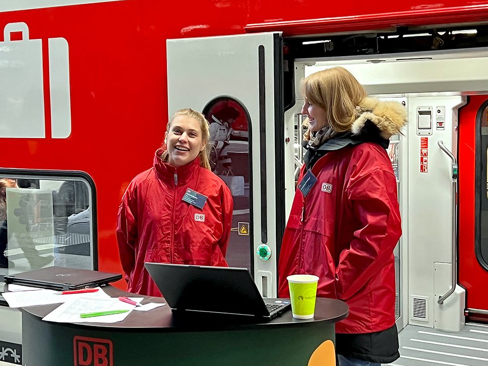 Zwei Mitarbeiterinnen stehen an einem Empfangspult vor dem Regionalzug