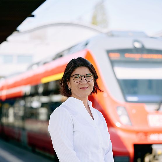 Eine Frau mit kurzen, dunklen Haaren und Brille lächelt in die Kamera, im Hintergrund ein roter Regionalzug.
