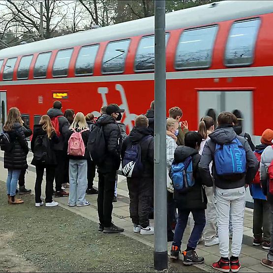 Eine Schulklasse steht am Bahnsteig. Ein roter Regionalzug fährt ein.