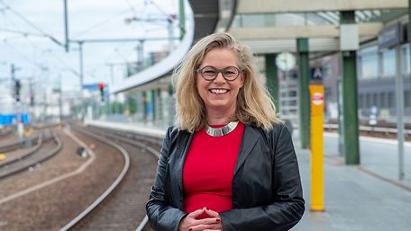 Eine blonde Frau mit Brille, rotem Shirt und schwarzer Jacke steht am Bahnsteig, im Hintergrund ein Bahnhof und Schienen.