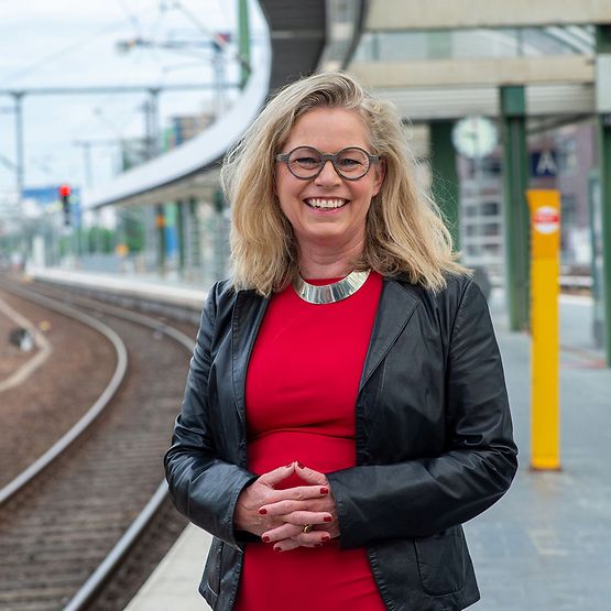 Eine blonde Frau mit Brille, rotem Shirt und schwarzer Jacke steht am Bahnsteig, im Hintergrund ein Bahnhof und Schienen.