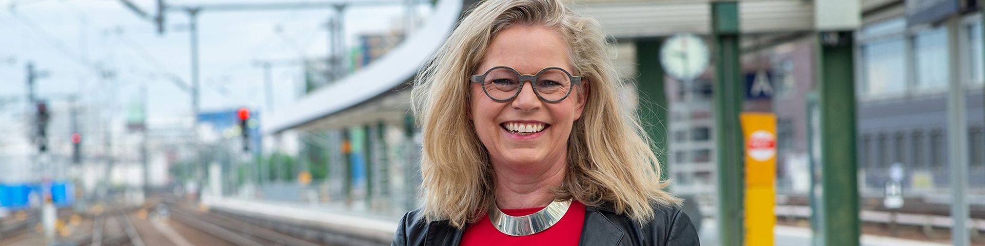Eine blonde Frau mit Brille, rotem Shirt und schwarzer Jacke steht am Bahnsteig, im Hintergrund ein Bahnhof und Schienen.