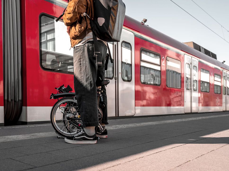 Eine Person steht am Bahnsteig, während ein roter Zug einfährt. Zwischen den Beinen steht das zusammengeklappte Faltrad.