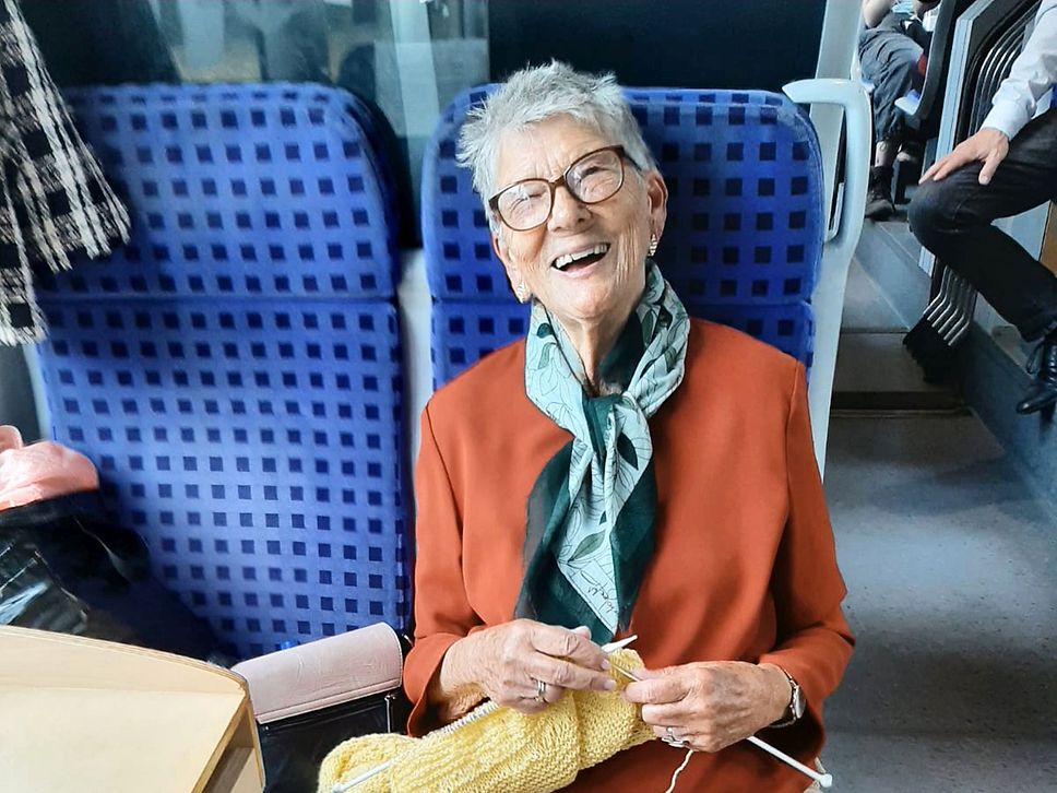 Eine ältere Dame mit rotem Pulli und grünem Schal sitzt in einem Regionalzug auf einem blauen Sitz.
