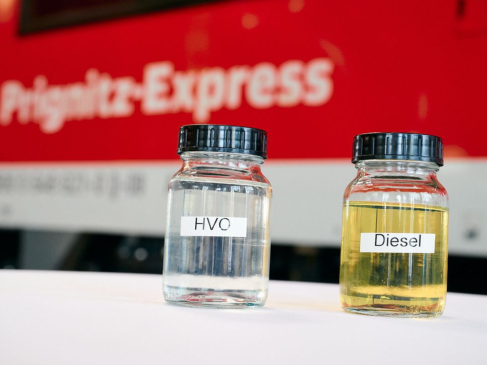 Zwei Schraubgläser mit Flüssigkeiten und den Beschriftungen HVO und Diesel stehen auf einem Tisch vor einem Regionalzug.