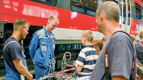 Zwei junge Männer erklären einem Jungen etwas, im Hintergrund ein roter Zug.