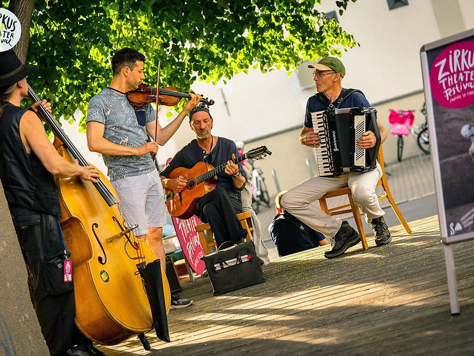 Eine vierköpfige Band spielt ein Lied mit Kontrabass, Geige, Gitarre und Akkordeon.