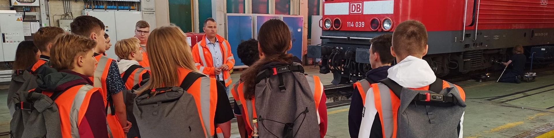 Mädchen und Jungs in Warnwesten und mit Rucksäcken sind von hinten zu sehen. Sie blicken auf einen roten Zug.