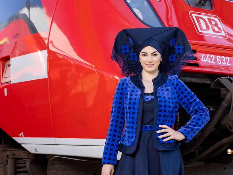 Eine Frau trägt die traditionelle Spreewald-Tracht mit DB-Stoff. Sie steht vor einem roten Regionalzug.