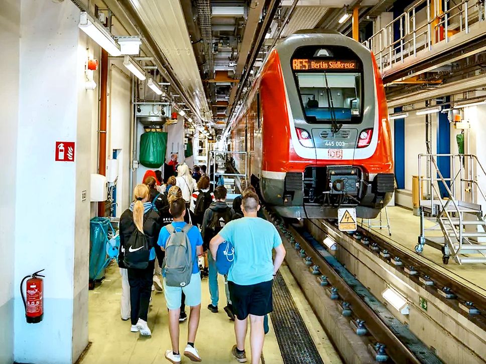 Links Schüler:innen, die in einem Werk laufen. Rechts ein roter Regionalzug über einem Werkstattgraben.