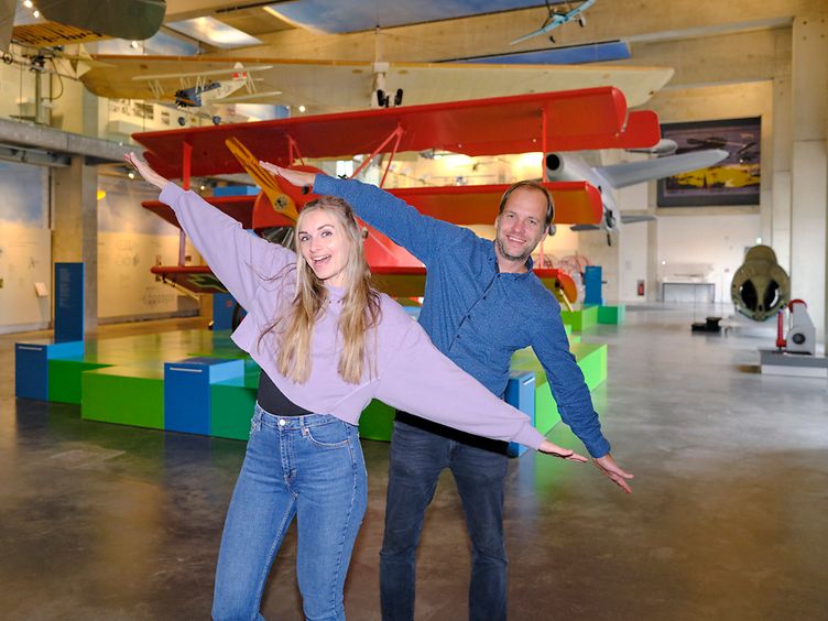 Maike und Olli posieren mit ausgestrecken Armen vor Flugzeugmodellen im Phantechnikum.