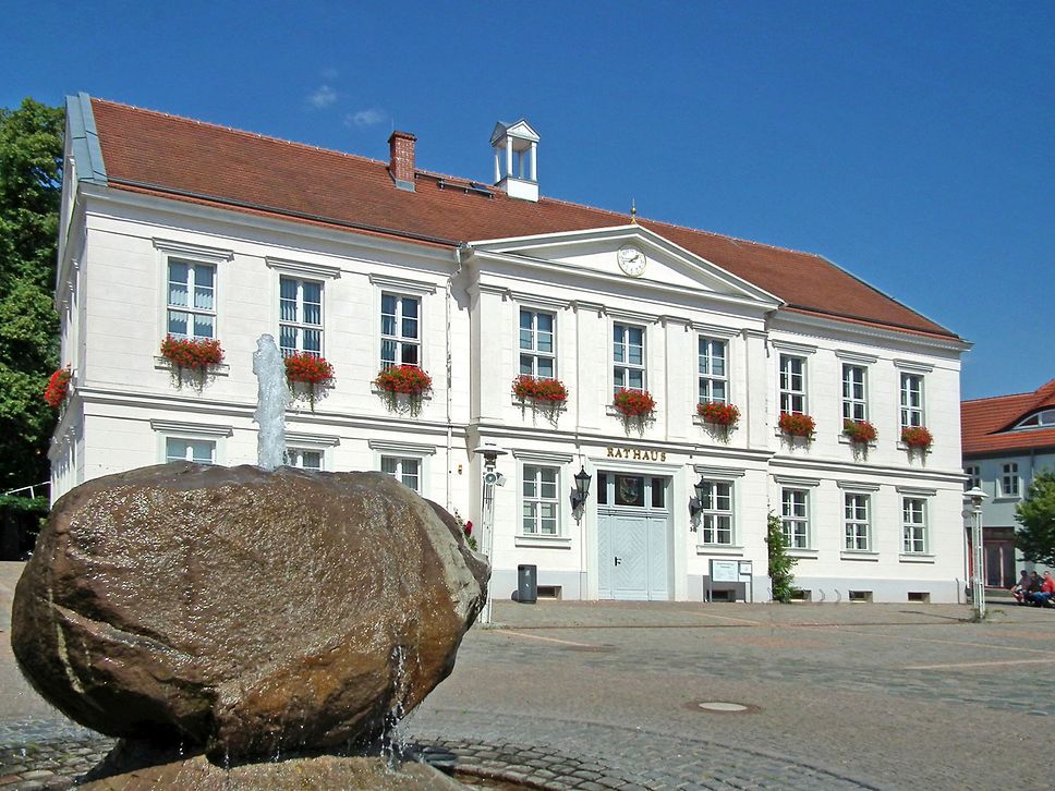 Rathaus in Pritzwalk, wießes Haus mit rotem Dach, links im Vordergrund ein Springbrunnen aus Stein.