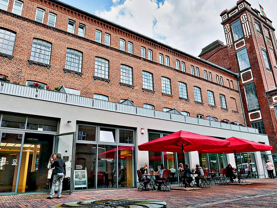 Außenansicht Museumsfabrik Pritzwalk. Gebäude mit rotem Klinkerstein, davor rote Schirme und Sitzmöglichkeiten.