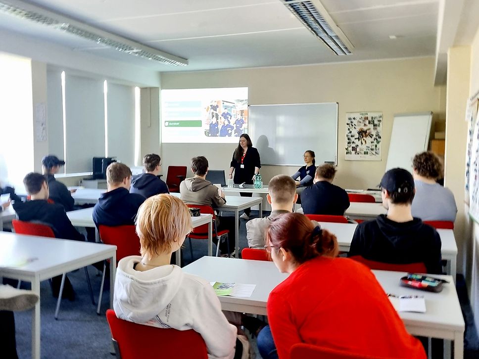 Blick in eine Klasse, Schüler:innen sind von hinten zu sehen, vorne referiert eine Frau.