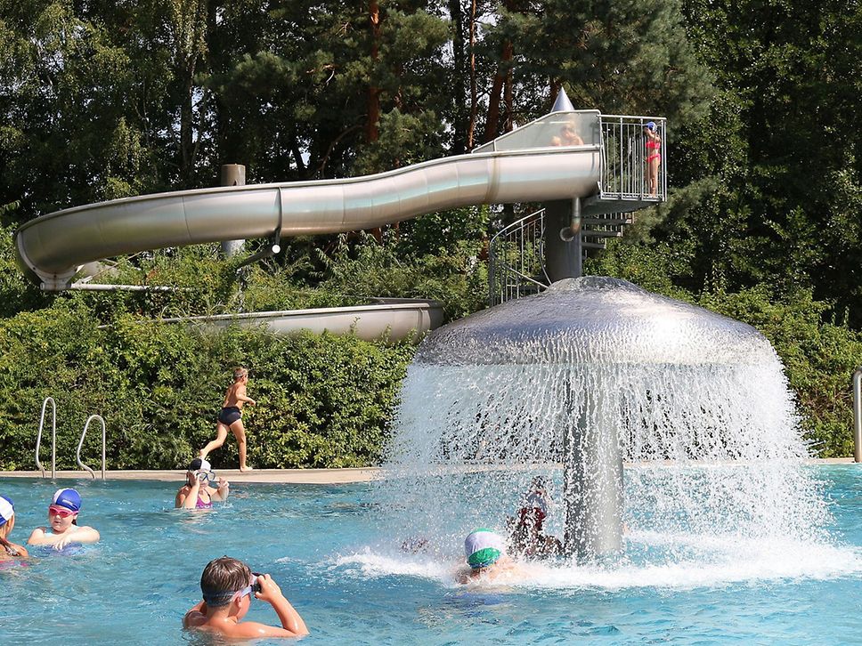 Blick ins Freibad, im Hintergrund eine Rutsche, mittig ein Wasserpilz.