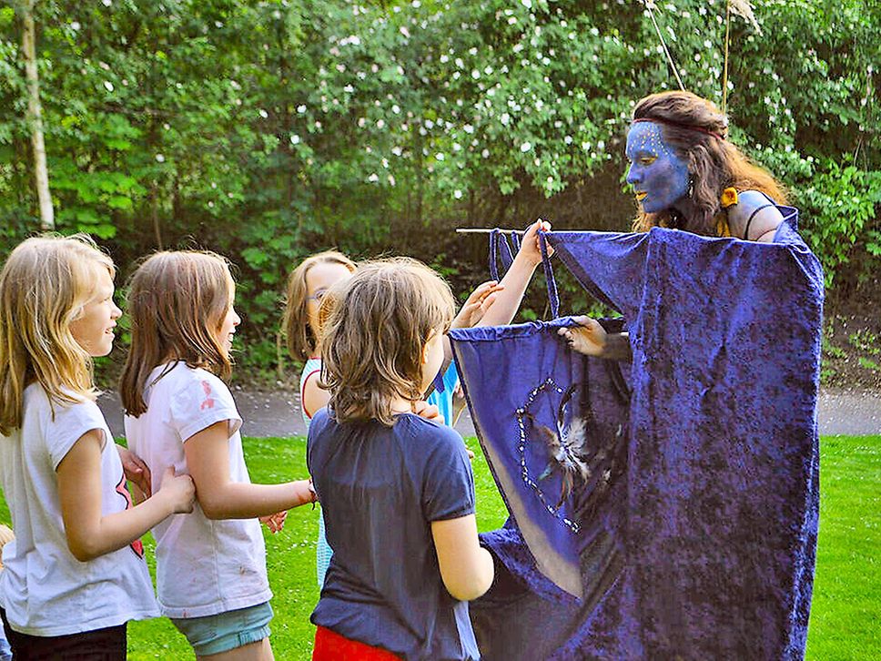 Eine in blauem Gewand und mit blauem Gesicht verkleidete Frau steht vor einer Gruppe Kinder.