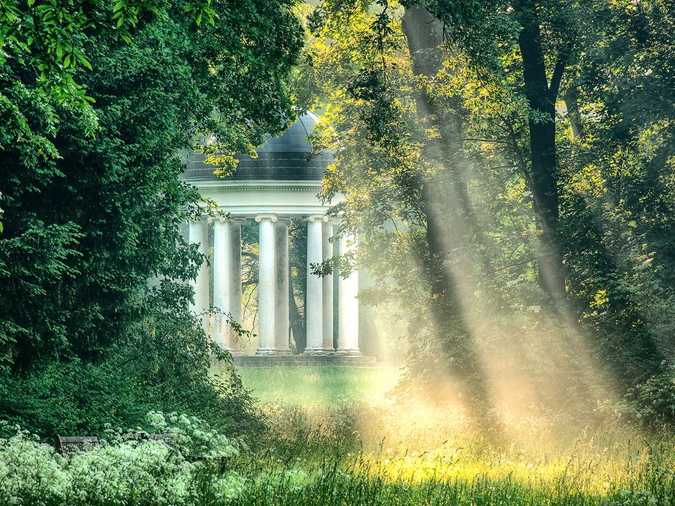 Weiße Säulen schimmern durch grüne Bäume hindurch, warme Sonnenstrahlen beleuchten den Park