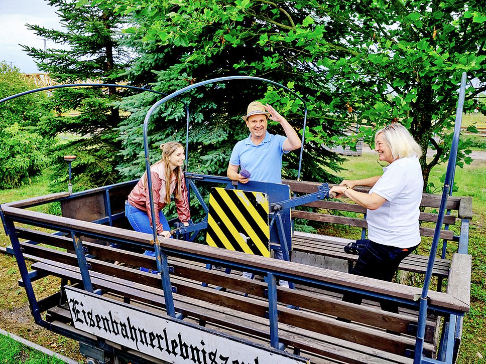 Drei Personen stehen in einer Draisine, zwei Frauen betätigen lachend den Handhebel, der Mann in der Mitte grüßt in die Kamera