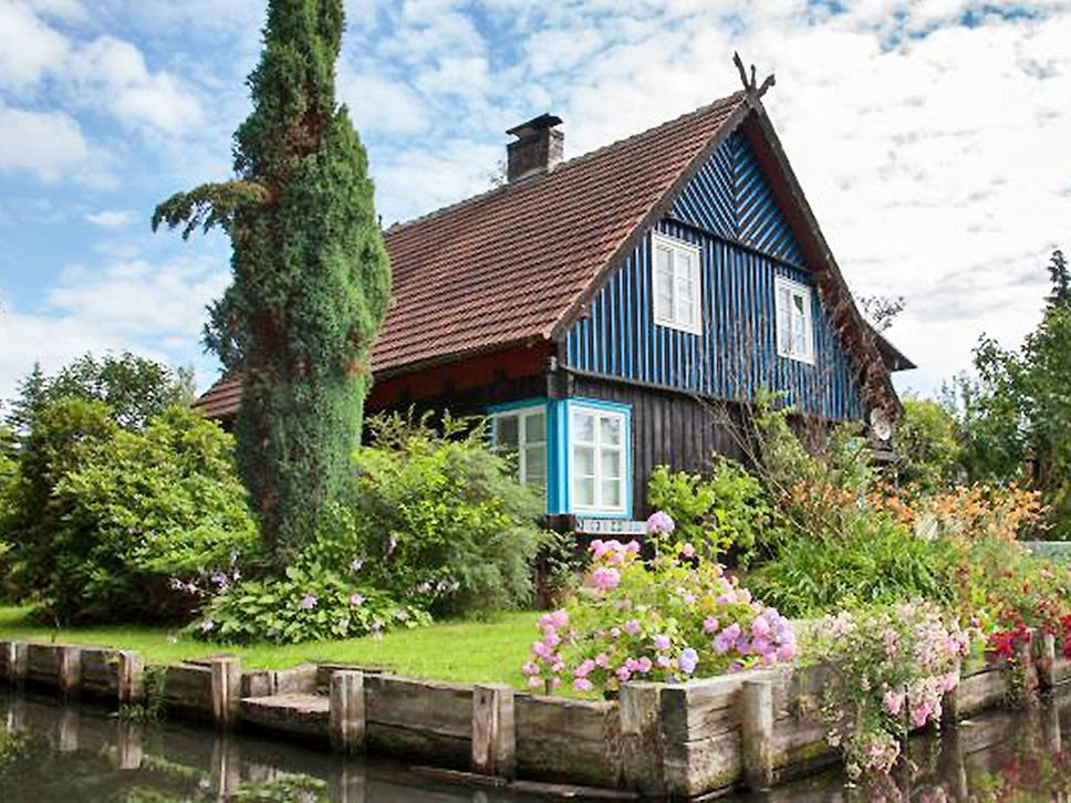 Foto eines blauen Blockbohlenhauses mit rotem Ziegeldach umgeben von grünem Garten und Wasser.