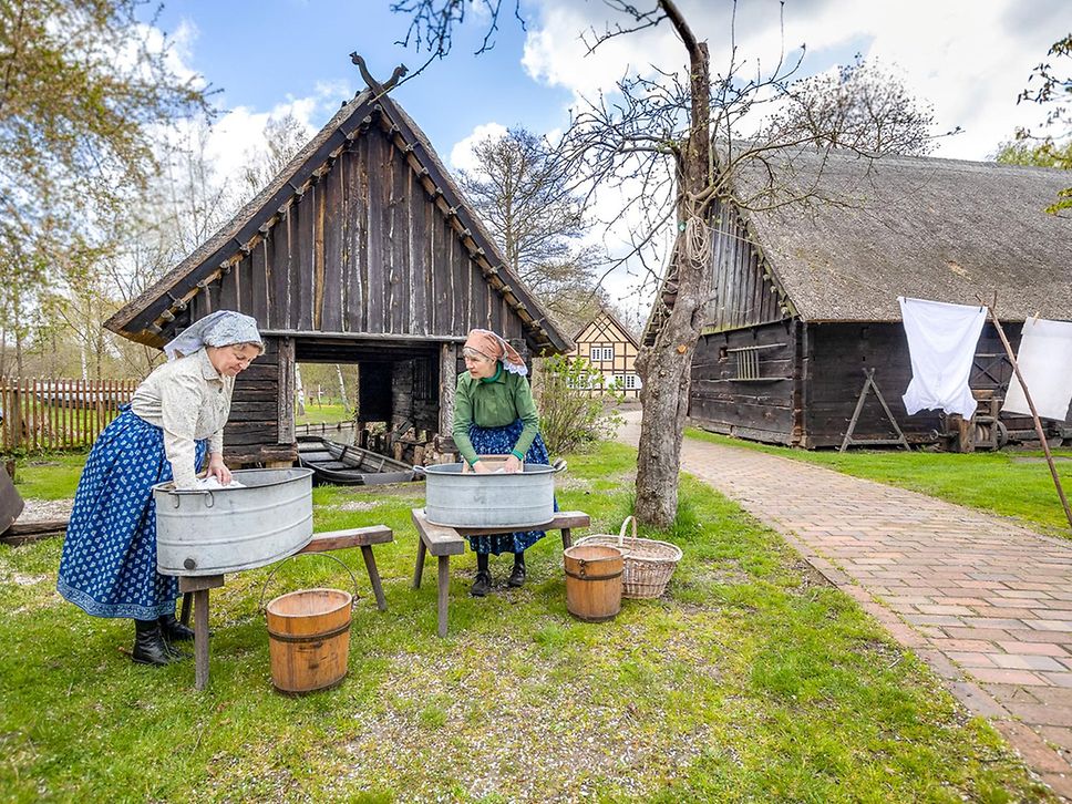 Zwei Frauen in altertümlicher Kleidung und mit Kopftuch stehen an einem Waschplatz und waschen in einer Zinkwanne.