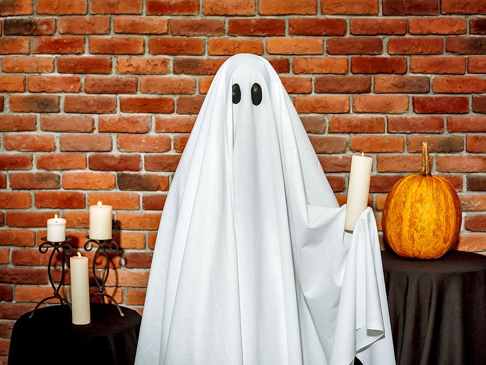 Eine Person als Gespenst verkleidet mit Kerze in der Hand, im Hintergrund ein Kürbis und weitere Kerzen.