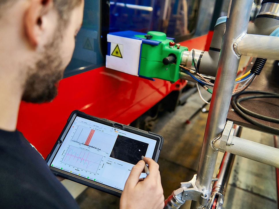 Blick über die Schulter eines Mannes mit Bart, er schaut auf ein Tablet, vor ihm der Laser an der Scheibe.