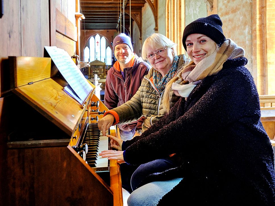 Zwei Frauen und ein Mann sitzen an einer Orgel und lächeln in die Kamera.