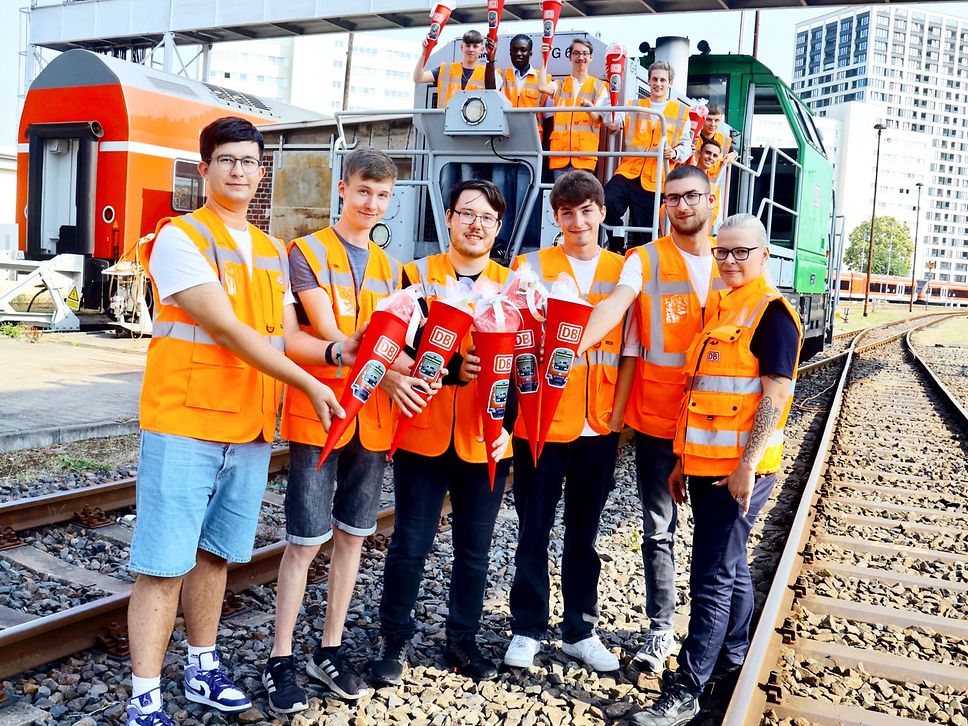 Eine Gruppe in orangefarbenen Warnwesten steht vor einer Lok und hält rote Zuckertüten in der Hand.