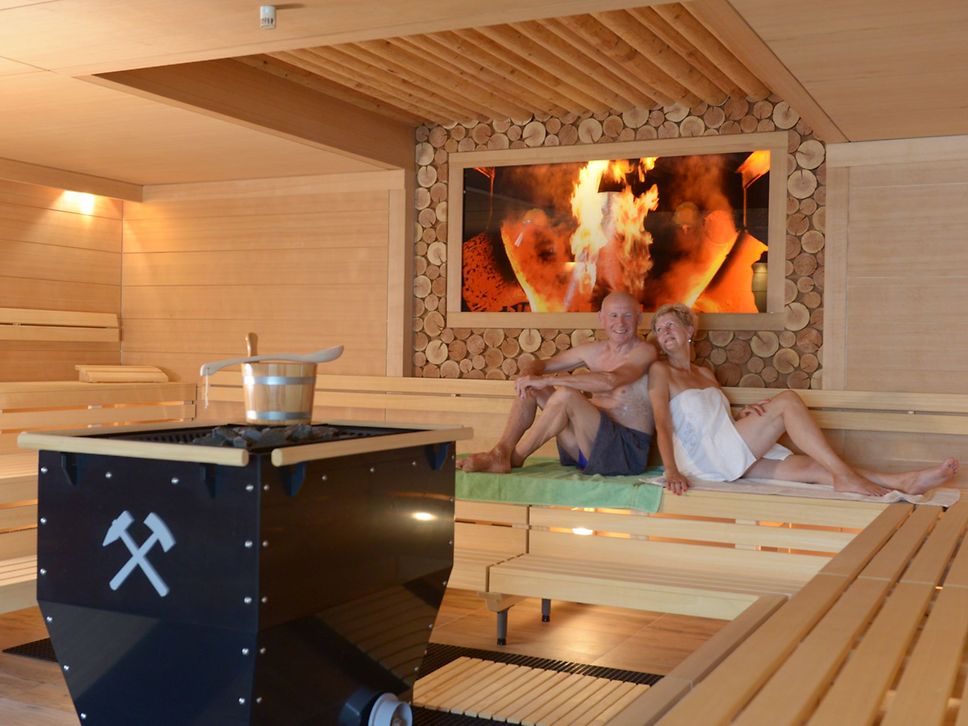 Ein Paar sitzt in einer Sauna, in der Mitte ein Bergwerkswagen, der zum Ofen umfunktioniert wurde.