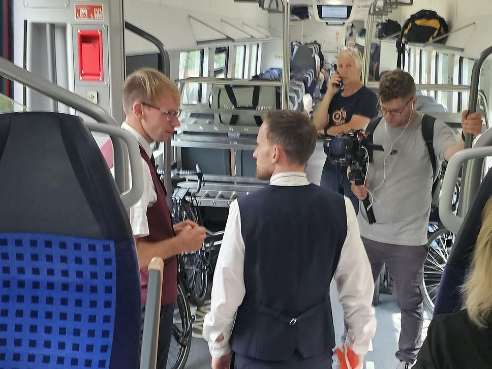 Gruppe junger Männer im Zug, einer filmt zwei andere im Gespräch.