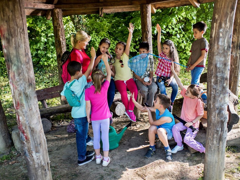 Eine Gruppe von Schüler:innen sitzt unter einem Unterstand im Wald, Kinder melden sich.