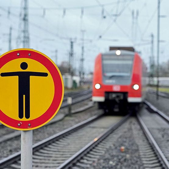 Im Hintergrund ein roter Zug auf Schienen, im Vordergrund ein "Betreten verboten"-Schild.