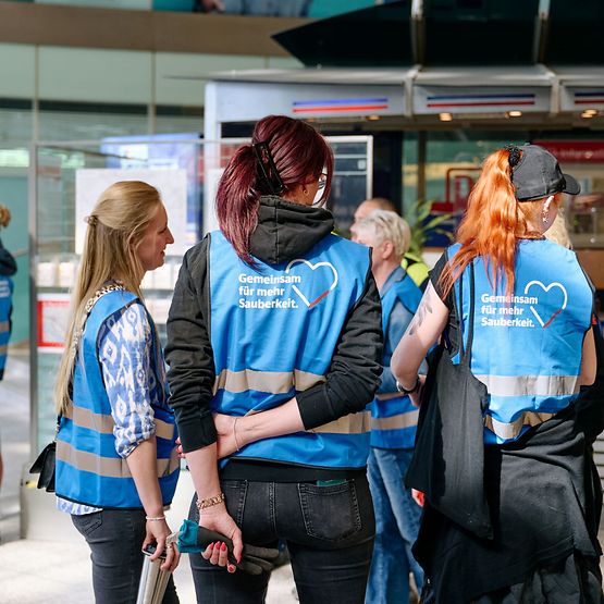 Rückansicht von DB-Mitarbeitenden in blauen Westen mit Aufschrift "Gemeinsam für mehr Sauberkeit".
