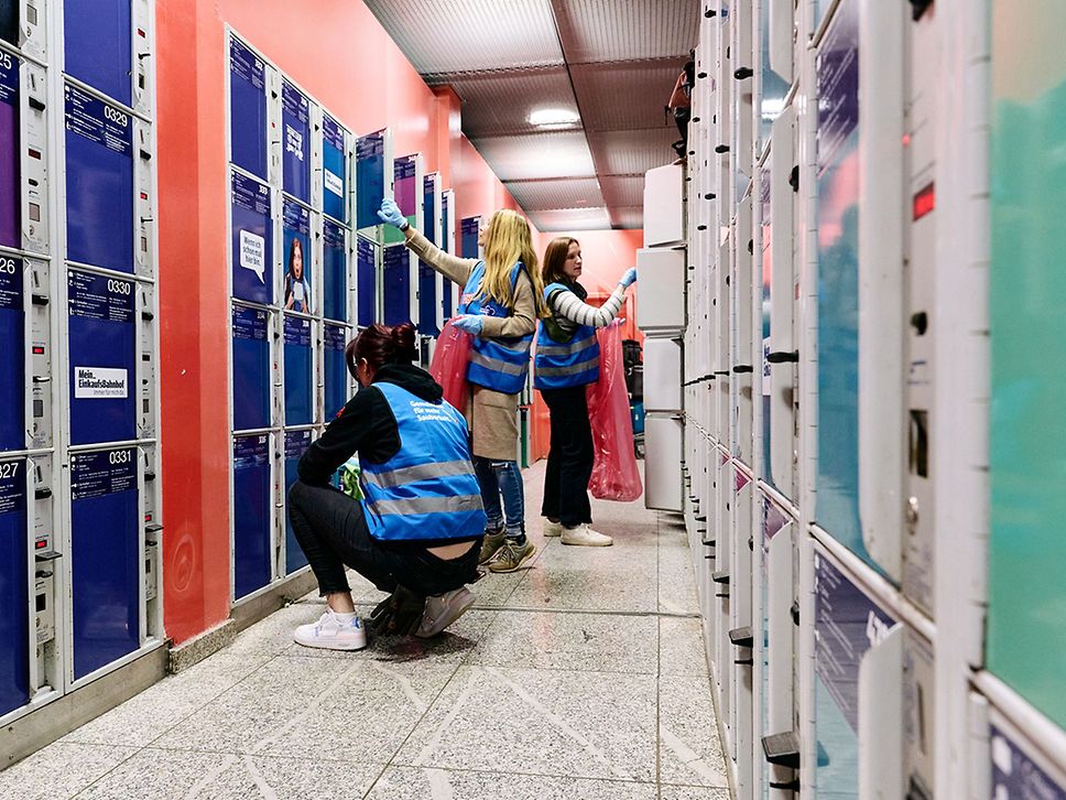 Drei Frauen mit Reinigungsutensilien säubern eine Reihe Schließfächer im Bahnhof.