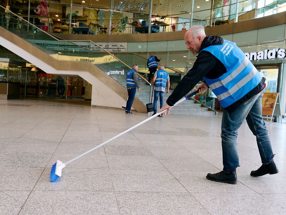 Ein Mann in Jeans, schwarzem Pulli und blauer Warnweste kehrt mit einem Besen die Empfangshalle im Ostbahnhof.