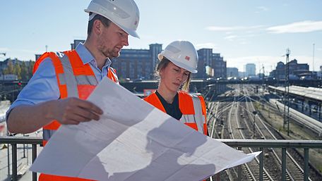 Zwei Bahn-Ingenieure in orangenen Westenschauen auf Baupläne.