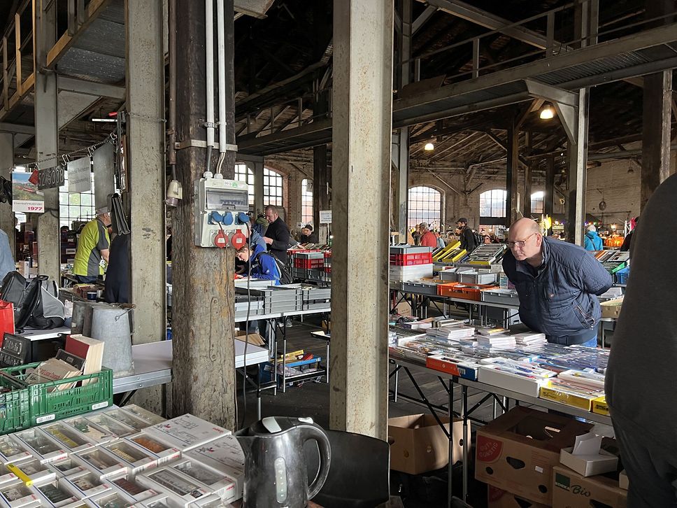 Blick in eine Halle mit vielen Verkaufsständen an denen Besucher:innen sich umschauen.