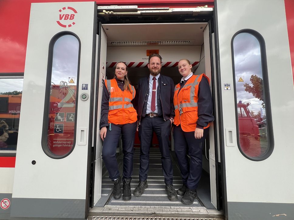 Drei Mitarbeitende von DB Regio Nordost stehen in der offenen Tür eines roten Regionalzugs und lächeln in die Kamera.
