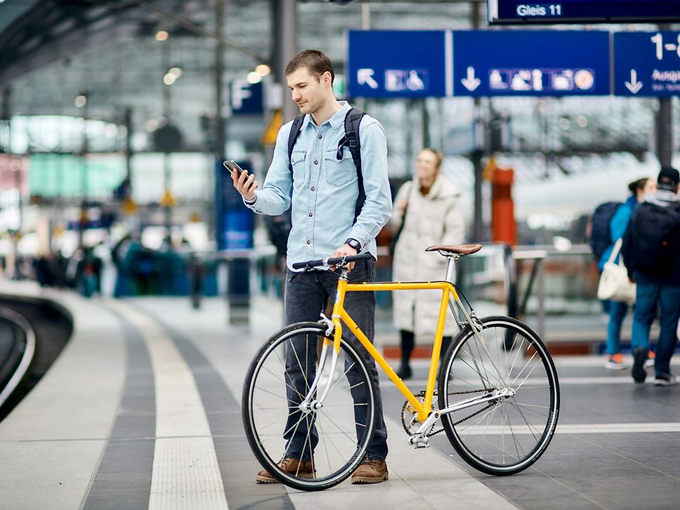 Ein Fahrgast steht mit seinem gelben Fahrrad am Bahnsteig und schaut aufs Handy.