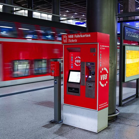 Ein roter VBB-Automat für Fahrausweise steht auf einem Bahnsteig, im Hintergrund ein roter Regionalzug.