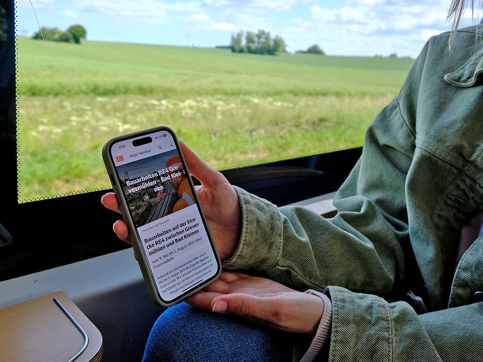 Jemand sitzt im Zug und zeigt sein Smartphone mit in die Kamera.