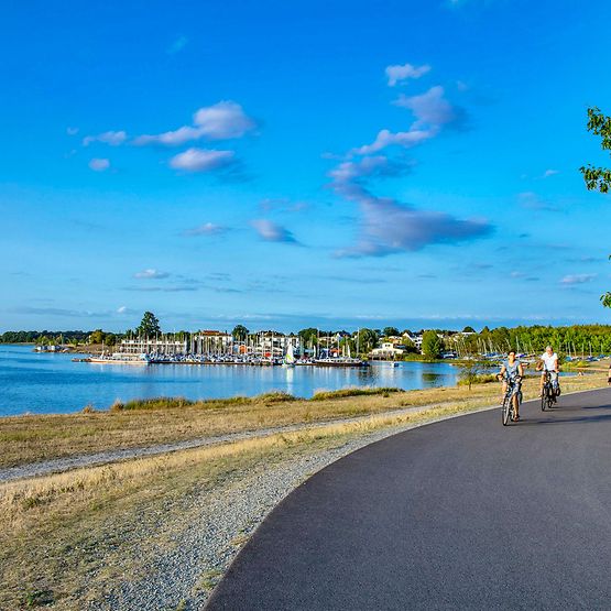 Weg um den Cospudener See, im Hintergrund der Hafen, zwei Menschen fahren Fahrrad.