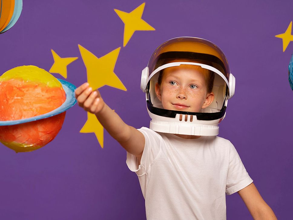 Ein Junge trägt einen Astronautenhelm und greift nach einem selbst gebastelten Planeten.