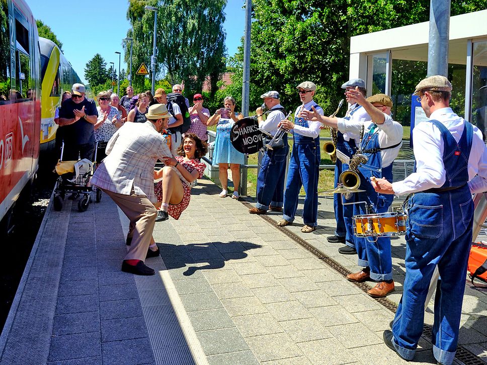 Auf einem Bahnsteig stehen feiernde Menschen, ein Pärchen tanzt Swing im Stil der 20er Jahre, daneben spielt eine Musikkapelle.