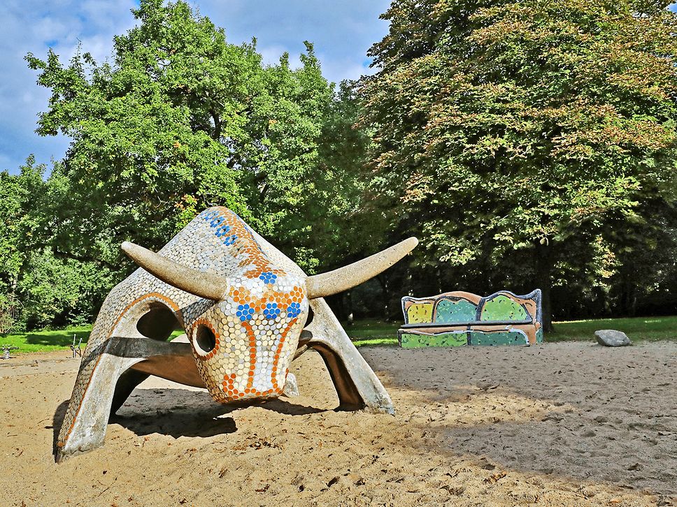 Blick auf eine bunte Skulptur, die aussieht wie ein Stier. Im Hintergrund grüne Bäume. Die Figur steht auf Sand.