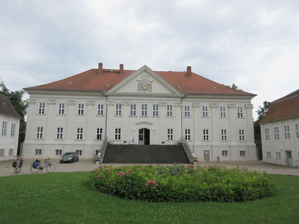 Blick auf ein herrschaftliches Haus mit vielen Fenstern und rotem Dach und breiter Treppe vor dem Eingang, davor Grünfläche.