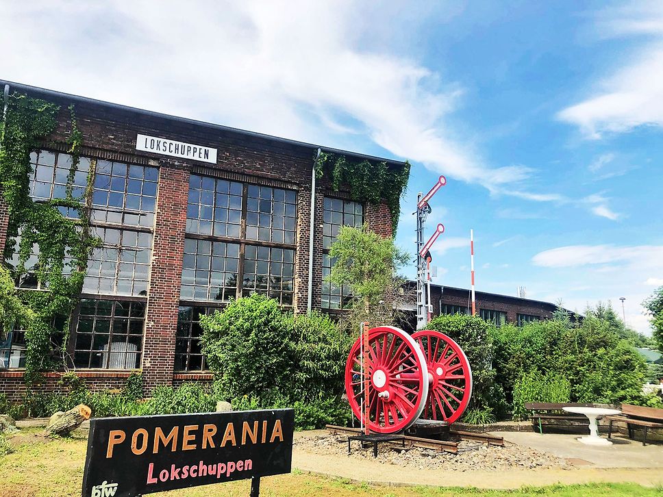 Blick auf den Eingang des Lokschuppens Pasewalk, im Hintergrund ein großes Wagenrad in rot.
